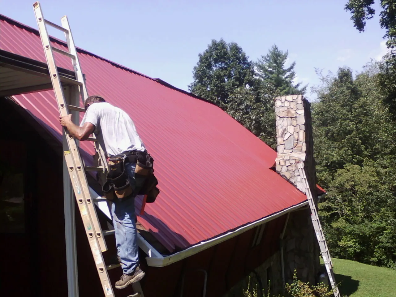 Roofer on a ladder beside a red metal roof and stone chimney — Soffit & Fascia Repair by Roofing Brothers in Edenton